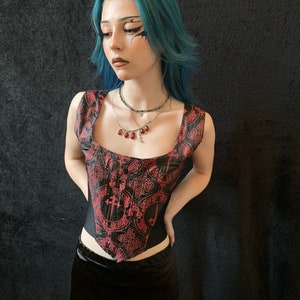 Op de afbeelding: Een zwart en rood korset met een Keltisch knoop- en kruispatroon. Het korset heeft een vierkante halslijn en is mouwloos. Het model draagt een zwarte rok en heeft blauw haar.
