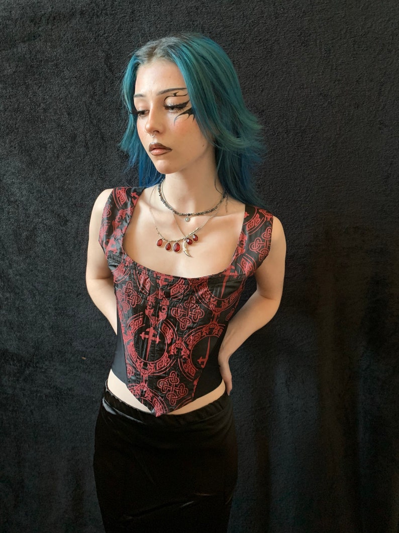 Op de afbeelding: Een zwart en rood korset met een Keltisch knoop- en kruispatroon. Het korset heeft een vierkante halslijn en is mouwloos. Het model draagt een zwarte rok en heeft blauw haar.