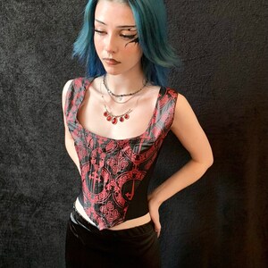 Op de afbeelding: Een persoon draagt ​​een zwart korset met een rood-zwart patroon. Het korsett heeft een vierkante halslijn en is mouwloos. De persoon draagt ​​ook een zilveren ketting met een rode edelsteen hanger.