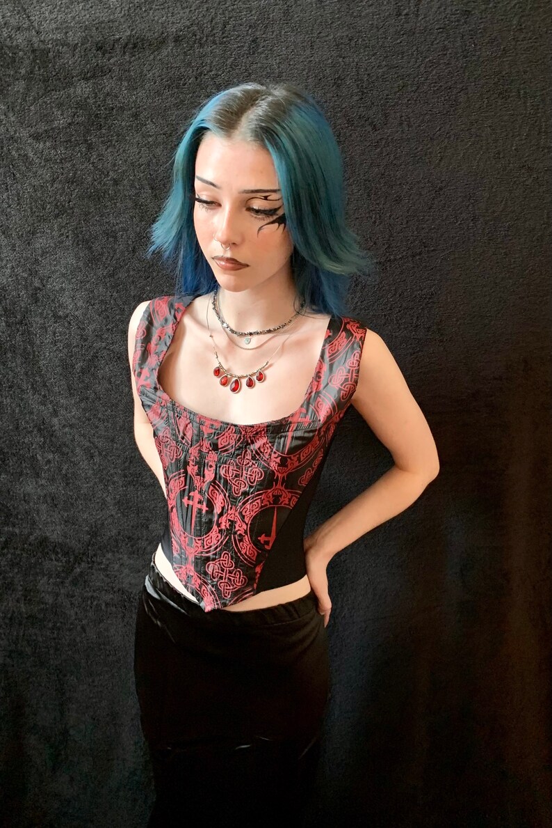 Op de afbeelding: Een persoon draagt ​​een zwart korset met een rood-zwart patroon. Het korsett heeft een vierkante halslijn en is mouwloos. De persoon draagt ​​ook een zilveren ketting met een rode edelsteen hanger.