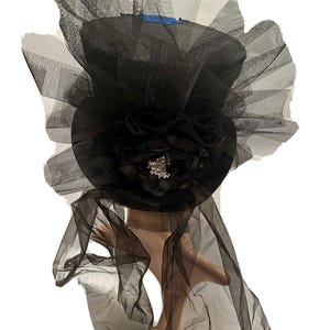 May include: Black top hat with a large black flower and a silver-colored embellishment. The hat is adorned with layers of black tulle, creating a dramatic and elegant look. A blue ribbon is visible on the top of the hat.