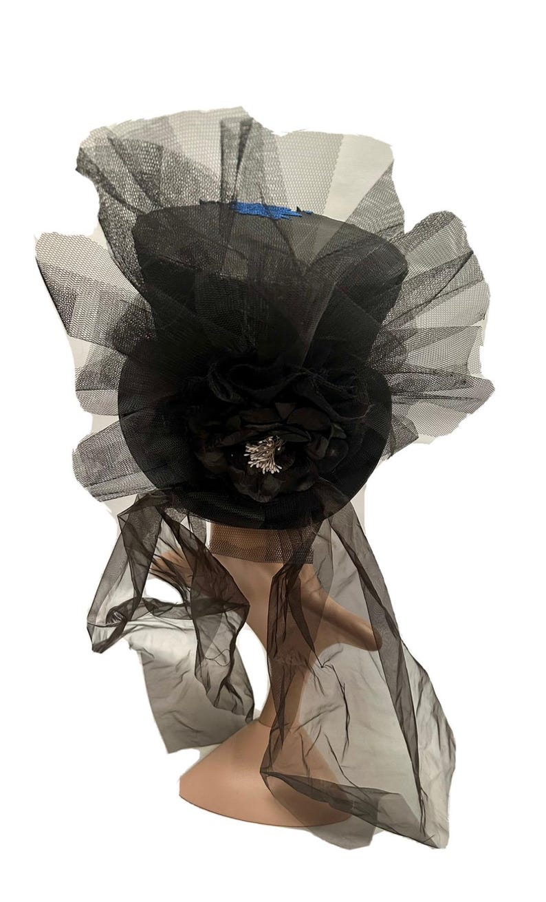 May include: Black top hat with a large black flower and a silver-colored embellishment. The hat is adorned with layers of black tulle, creating a dramatic and elegant look. A blue ribbon is visible on the top of the hat.
