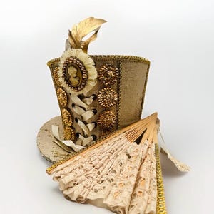 May include: A decorative steampunk-style top hat with gold embellishments, a cameo, and a gold feather. A matching lace fan rests beside the hat. The hat features a corset-style ribbon design and is decorated with gold sequins and floral accents.