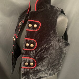May include: A black velvet vest with a red trim and silver buttons. The vest has a stand-up collar and a pointed hem.