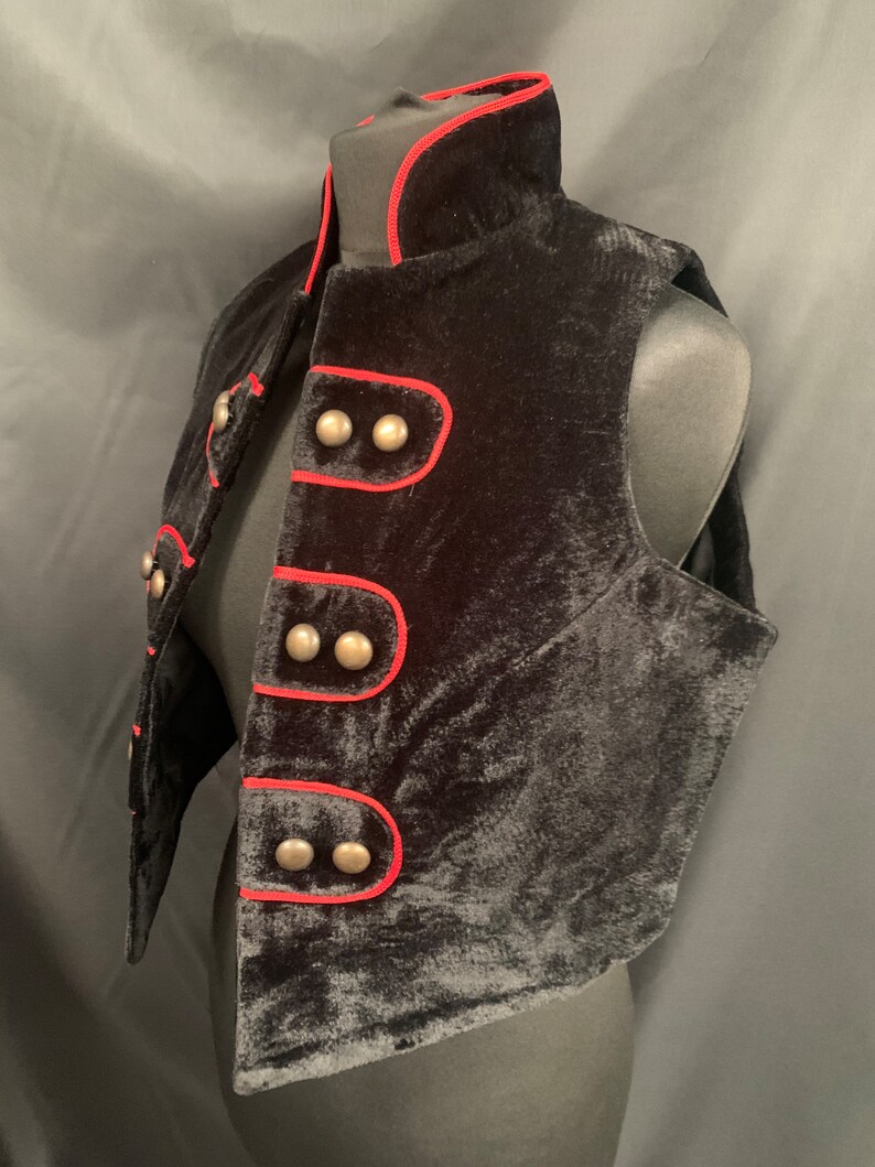 May include: A black velvet vest with a red trim and silver buttons. The vest has a stand-up collar and a pointed hem.