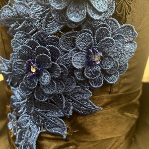 May include: Close-up of a dark blue fabric adorned with intricate, embroidered floral designs. The flowers are a vibrant shade of blue, with delicate detailing and sparkling embellishments. The fabric has a satin-like sheen, adding to the elegance of the piece.