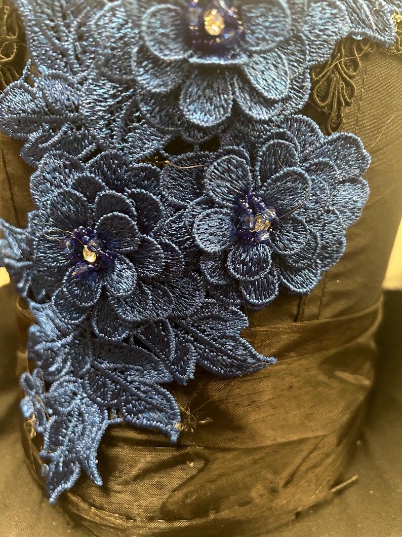 May include: Close-up of a dark blue fabric adorned with intricate, embroidered floral designs. The flowers are a vibrant shade of blue, with delicate detailing and sparkling embellishments. The fabric has a satin-like sheen, adding to the elegance of the piece.