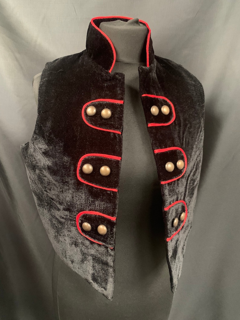 May include: A black velvet vest with a red trim and silver buttons. The vest has a mandarin collar and a cropped length.
