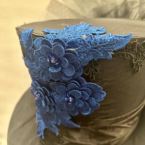 May include: A black top hat with a dark blue satin band and a decorative blue floral lace design. The hat features a dark blue floral lace design with small, sparkling embellishments.