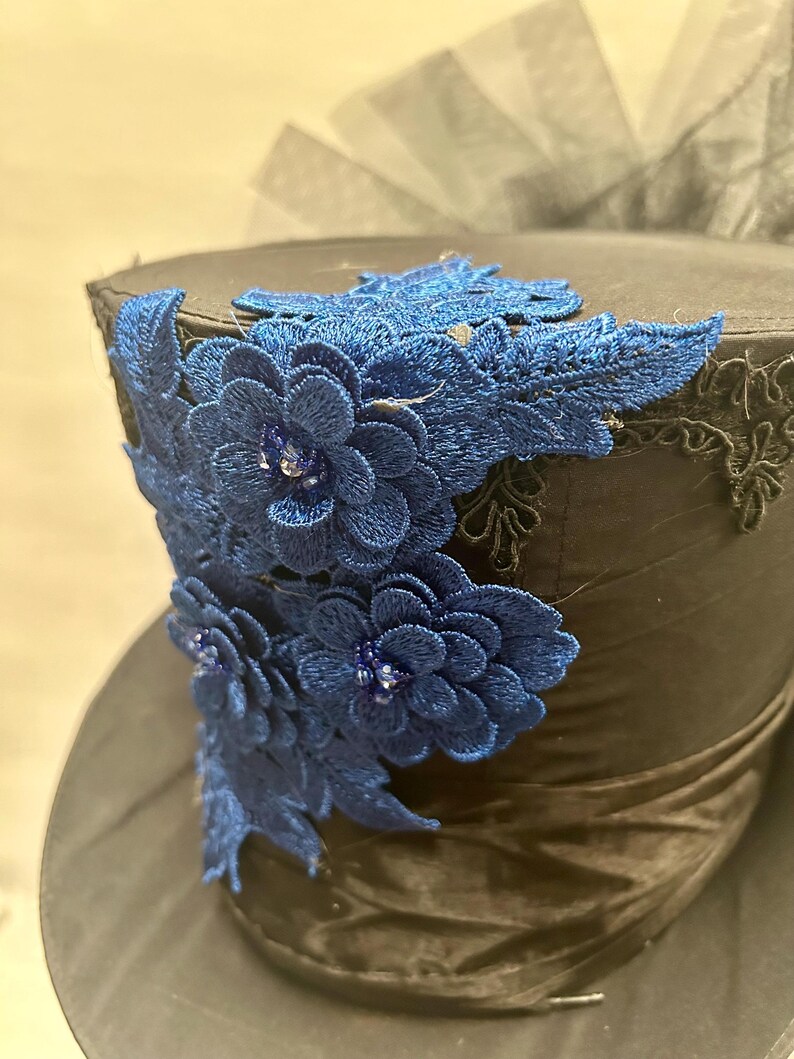 May include: A black top hat with a dark blue satin band and a decorative blue floral lace design. The hat features a dark blue floral lace design with small, sparkling embellishments.