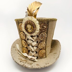 May include: A tan sequined top hat with gold embellishments, a cameo, and a gold feather. The hat features a cream-coloured ribbon and decorative gold accents. The hat is designed in a steampunk or Victorian style.