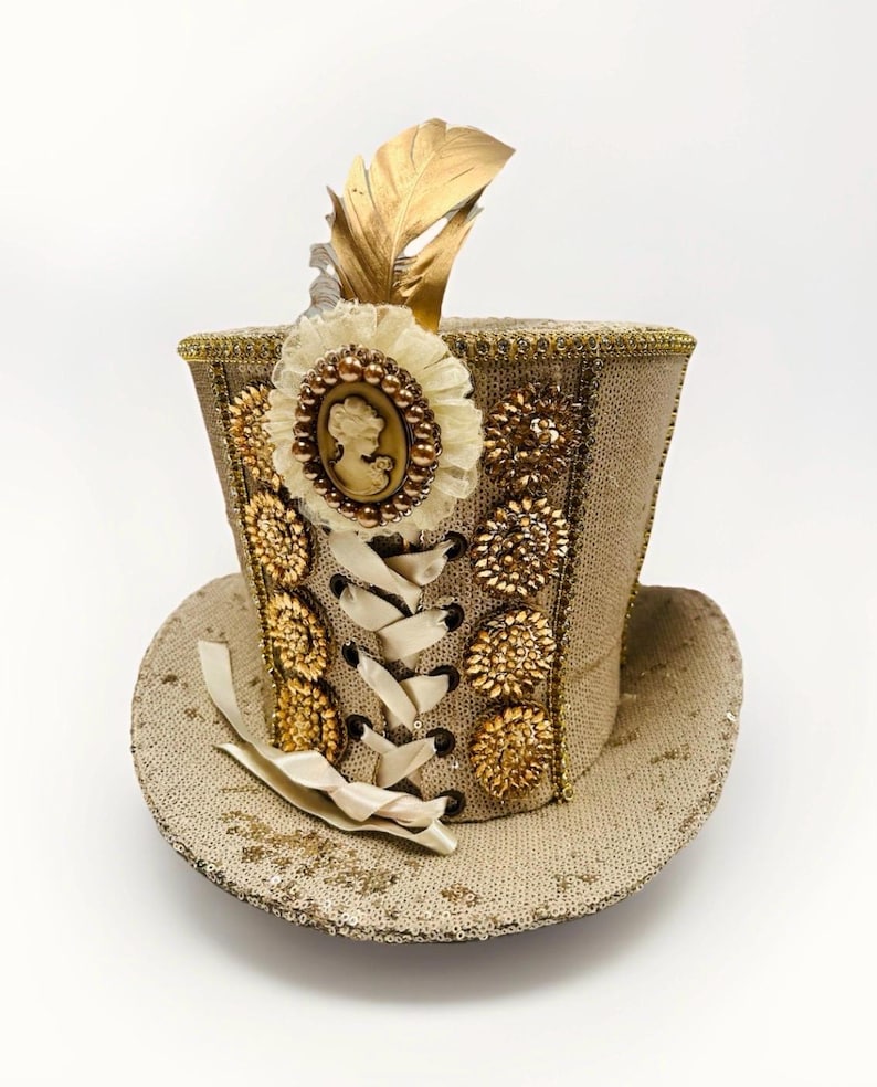 May include: A tan sequined top hat with gold embellishments, a cameo, and a gold feather. The hat features a cream-coloured ribbon and decorative gold accents. The hat is designed in a steampunk or Victorian style.