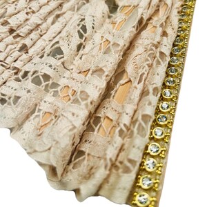 May include: A beige lace fan with a decorative gold trim. The fan has an intricate openwork design and is accented with sparkling clear gemstones. The fan's wooden frame is visible through the lace.
