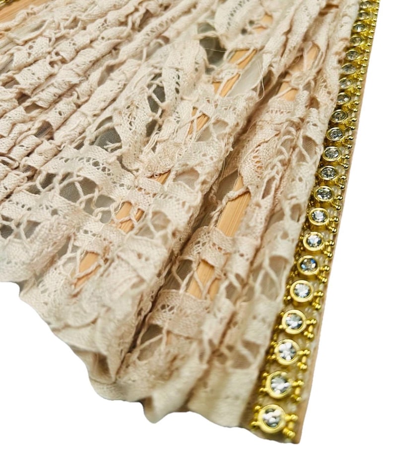 May include: A beige lace fan with a decorative gold trim. The fan has an intricate openwork design and is accented with sparkling clear gemstones. The fan's wooden frame is visible through the lace.