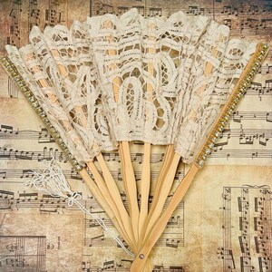 May include: Several open, cream-coloured lace fans with wooden handles. The edges are decorated with gold trim and rhinestones. A white tassel hangs from the base of one fan. The fans are displayed on a background of vintage sheet music.