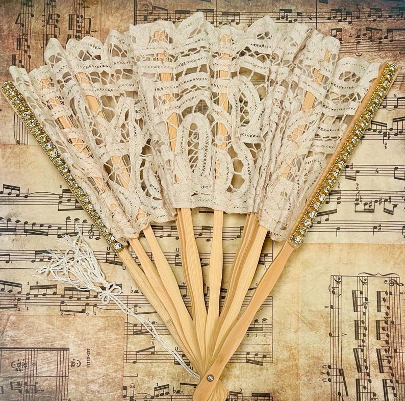 May include: Several open, cream-coloured lace fans with wooden handles. The edges are decorated with gold trim and rhinestones. A white tassel hangs from the base of one fan. The fans are displayed on a background of vintage sheet music.