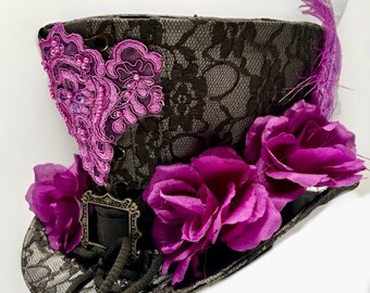 Può includere: Un cappello a cilindro nero con una sovrapposizione di pizzo, adornato con vivaci fiori viola e un'applicazione di pizzo viola decorativa. Una piccola cornice ornata e una piuma viola aggiungono ulteriore abbellimento a questo cappello unico.