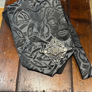 Puede incluir: Una corbata negra y gris estampada con una broche plateada. La corbata presenta un patrón repetitivo de caras y formas abstractas. La broche es adornada con un diseño ovalado central.