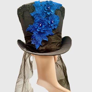 May include: Black top hat with a wide brim, adorned with blue floral lace and a black ribbon band. The hat features a sheer black veil and is a unique accessory for themed events or costumes.