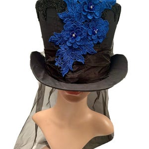 May include: Black top hat with a wide brim, adorned with blue floral lace and sparkling accents. A black sheer veil drapes from the hat. The hat is displayed on a mannequin head.