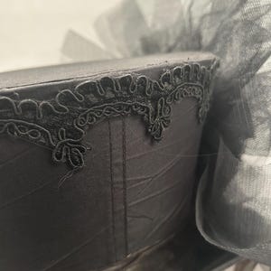 May include: A black hat with a decorative trim and a large bow made of sheer fabric. The hat's surface has a subtle sheen, and the trim features an intricate, floral-like design. The bow adds a touch of elegance to the overall design.