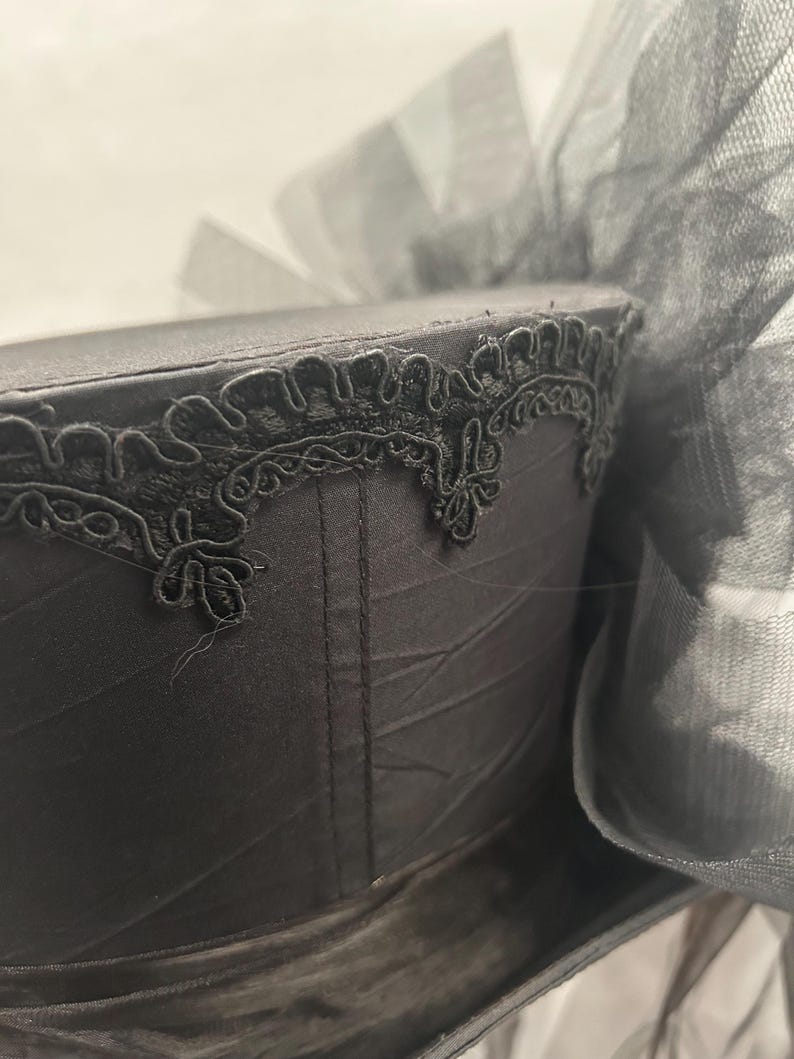 May include: A black hat with a decorative trim and a large bow made of sheer fabric. The hat's surface has a subtle sheen, and the trim features an intricate, floral-like design. The bow adds a touch of elegance to the overall design.