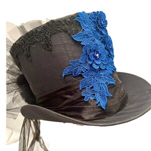 May include: Black top hat with a wide brim, adorned with a blue floral lace applique. The hat features a black ribbon band and a black lace trim along the top edge. A sheer black fabric is attached to the back of the hat.