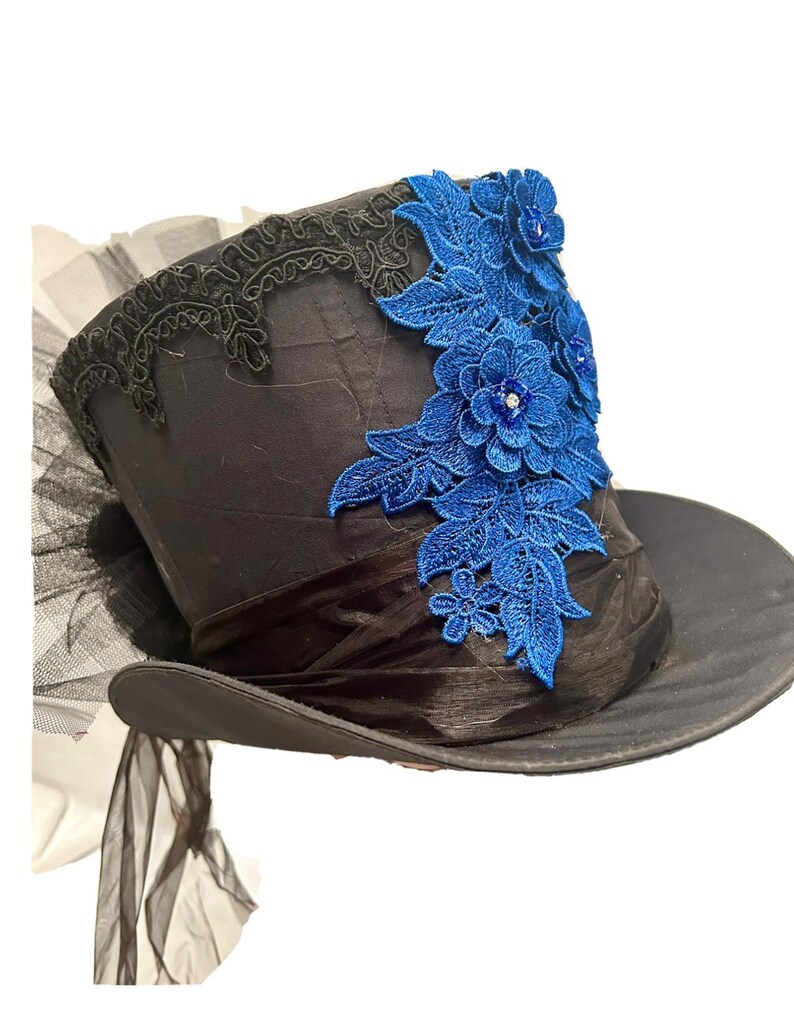May include: Black top hat with a wide brim, adorned with a blue floral lace applique. The hat features a black ribbon band and a black lace trim along the top edge. A sheer black fabric is attached to the back of the hat.