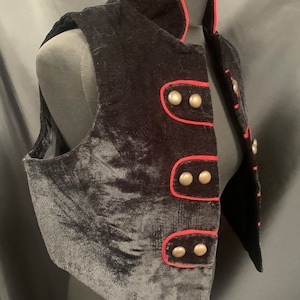 May include: A black velvet vest with a red trim and silver buttons. The vest has a stand-up collar and a fitted silhouette.