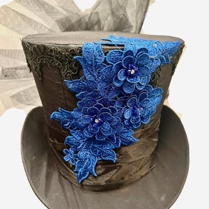 May include: A black top hat with a wide brim, adorned with a cluster of blue embroidered flowers and leaves. The hat features a black lace trim and a dark ribbon band. A black tulle fabric is visible in the background.