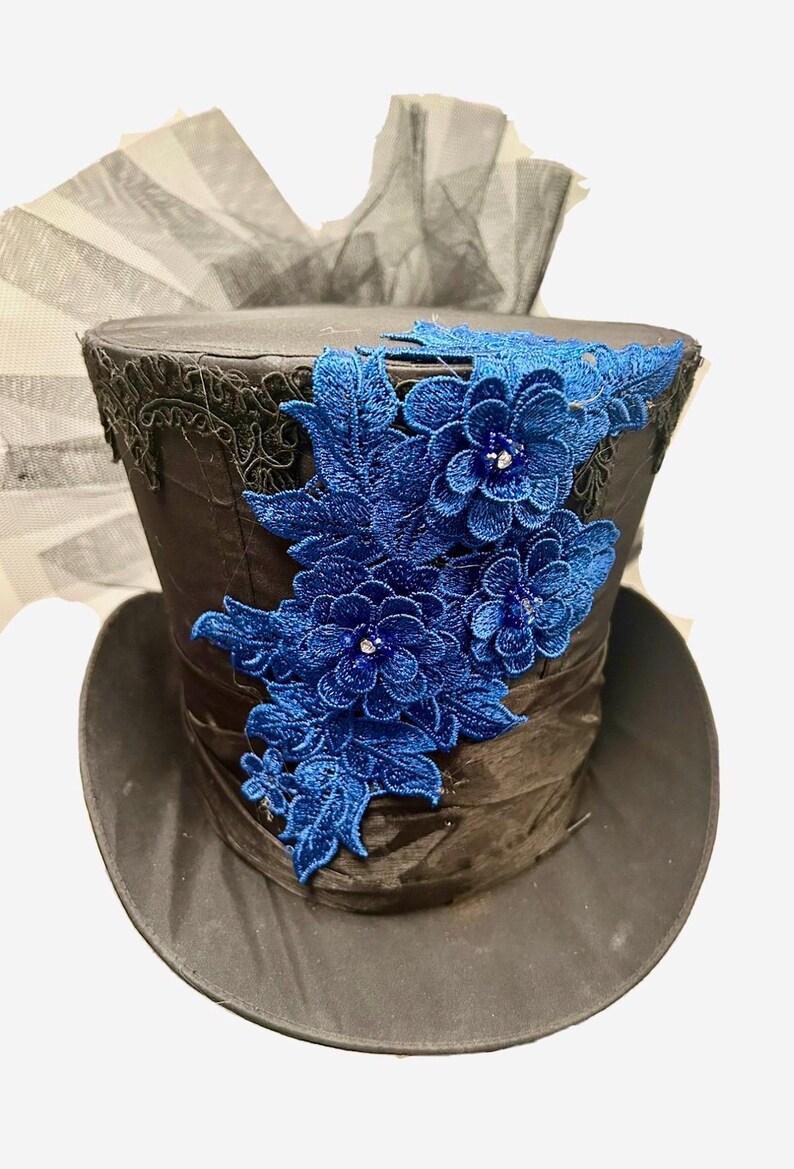 May include: A black top hat with a wide brim, adorned with a cluster of blue embroidered flowers and leaves. The hat features a black lace trim and a dark ribbon band. A black tulle fabric is visible in the background.