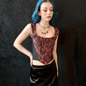 Op de afbeelding: Een zwart en rood korset met een geometrisch patroon en een zwarte satijnen rok. Het korset heeft een puntige halslijn en een gespeld rugpand. Het model draagt een zilveren ketting met rode kralen.