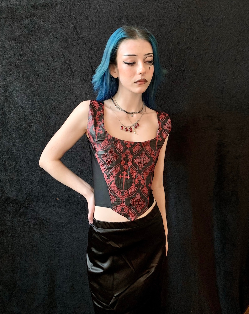 Op de afbeelding: Een zwart en rood korset met een geometrisch patroon en een zwarte satijnen rok. Het korset heeft een puntige halslijn en een gespeld rugpand. Het model draagt een zilveren ketting met rode kralen.
