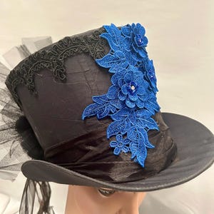 May include: Black top hat with a wide brim, adorned with a blue floral lace applique. The hat features a black ribbon band and a delicate black lace trim along the top edge. Black tulle and ribbons add a touch of elegance.