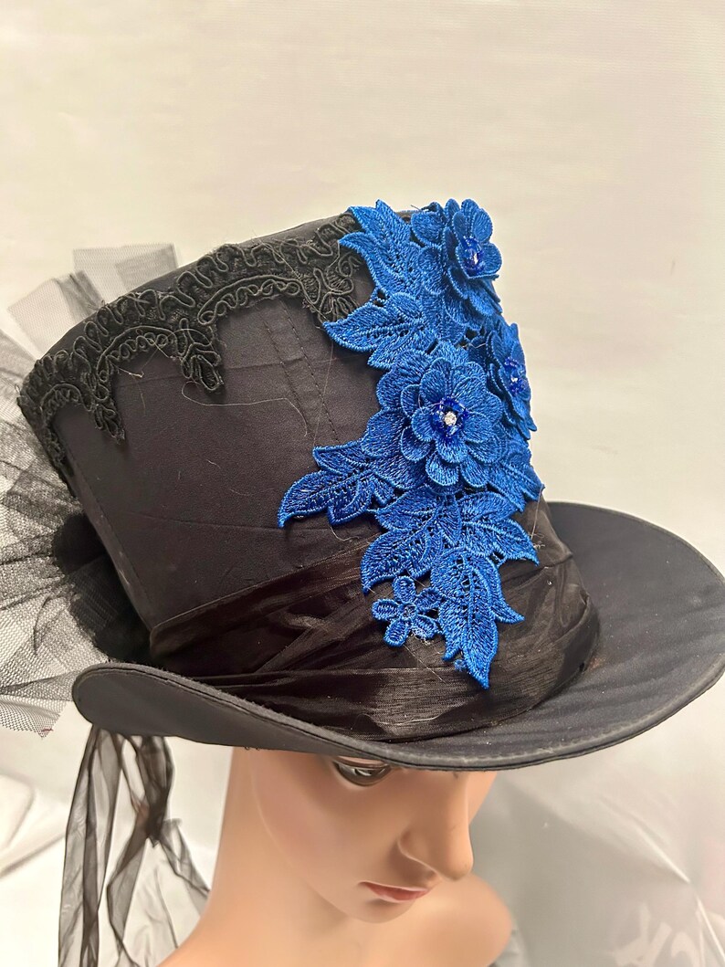 May include: Black top hat with a wide brim, adorned with a blue floral lace applique. The hat features a black ribbon band and a delicate black lace trim along the top edge. Black tulle and ribbons add a touch of elegance.