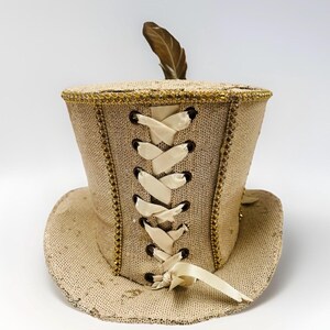 May include: A tan top hat with a wide brim, decorated with gold trim and sequins. The back has corset-style lacing with cream ribbons. A single brown feather is on top. The hat is likely for costume or theatrical use.