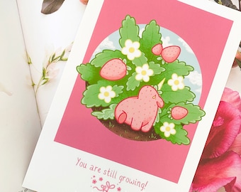 May include: A pink art print depicting a strawberry unicorn in a strawberry bush with white flowers. The print has a pink background and the text "You are still growing!" at the bottom. The print is on a white background.