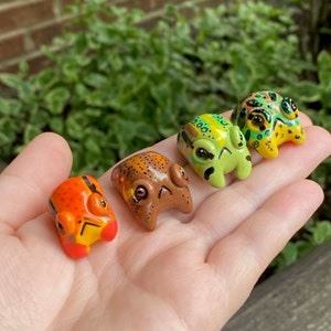 May include: Four colorful, handcrafted frog figurines in a variety of colors, including orange, brown, yellow, and green. Each frog has unique patterns and expressions, with visible eyes and detailed features. The frogs are small and appear to be made of clay or a similar material.