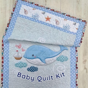 May include: A baby quilt kit featuring a blue whale, sailboat, and clouds. The quilt has a light blue background with a white border and a floral patterned edge. The text "Baby Quilt Kit" is displayed.