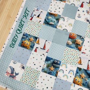 Puede incluir: Un kit de edredón para bebé con un diseño de patchwork con temas náuticos. El edredón incluye cuadrados con veleros, ballenas, estrellas de mar y arcoíris en tonos de azul, blanco y naranja. Las palabras "BABY QUILT KIT" están bordadas en verde.