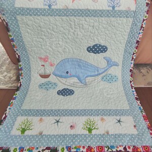 May include: A light blue and white quilt with a whale design. The quilt features a large blue whale, a sailboat, clouds, and sea-themed patterns. The border has a floral pattern.