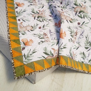 May include: A quilted blanket featuring a white background with a repeating pattern of deer, rabbits, and squirrels among green leaves and pinecones. The border consists of alternating orange and green triangles with a textured finish.