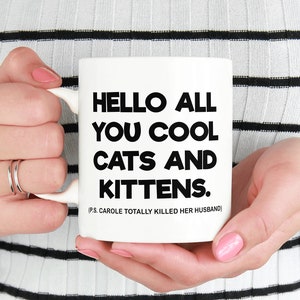 May include: White ceramic mug with black text that reads "Hello all you cool cats and kittens. PS. Carole totally killed her husband."
