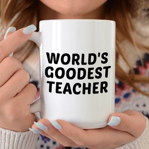 May include: A white ceramic mug with the text "WORLD'S GOODEST TEACHER" in bold black letters. The mug is held by a person wearing a patterned sweater and a white hooded jacket.