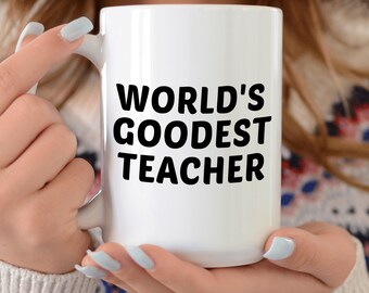 May include: A white ceramic mug with the text "WORLD'S GOODEST TEACHER" in bold black letters. The mug is held by a person wearing a patterned jumper and a white hooded jacket.