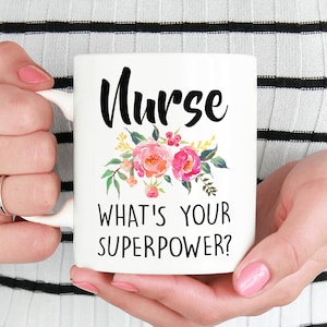 May include: White ceramic mug with the word "Nurse" in black script, adorned with a floral design in pink, orange, and green. The text "WHAT'S YOUR SUPERPOWER?" is below the floral arrangement. The mug is held by a person.