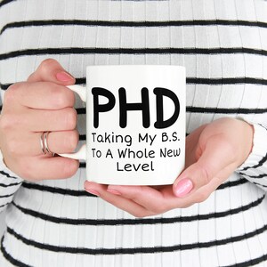 Puede incluir: Taza de cer&aacute;mica blanca con texto negro que dice "PHD Taking My B.S. To A Whole New Level"