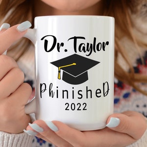May include: White ceramic mug with black text that reads "Dr. Taylor Phinished 2022" and a black graduation cap with a gold tassel.
