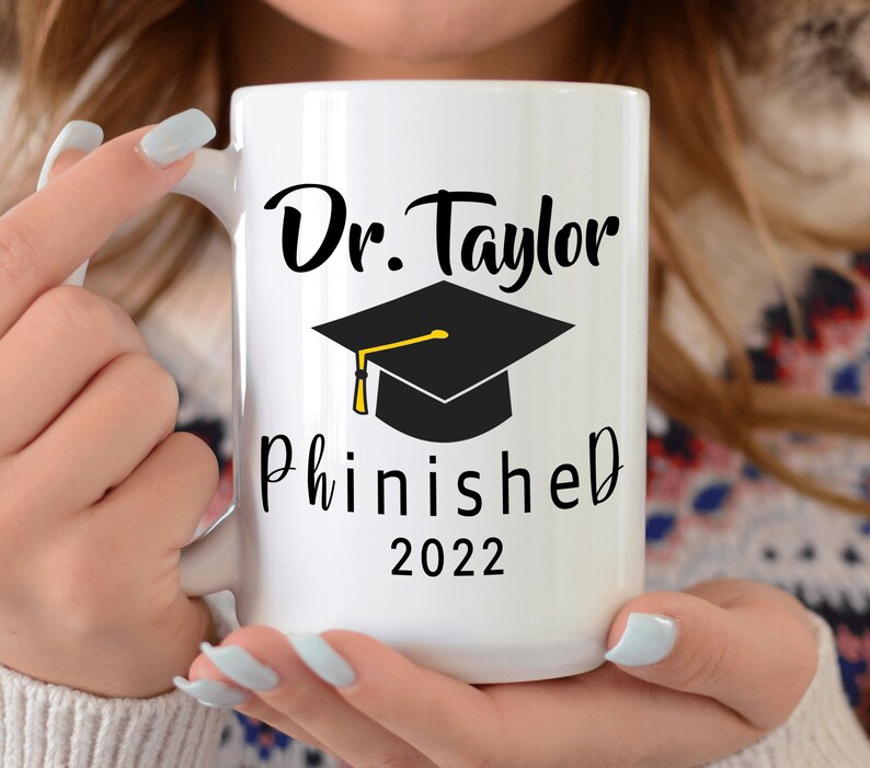 May include: White ceramic mug with black text that reads "Dr. Taylor Phinished 2022" and a black graduation cap with a gold tassel.