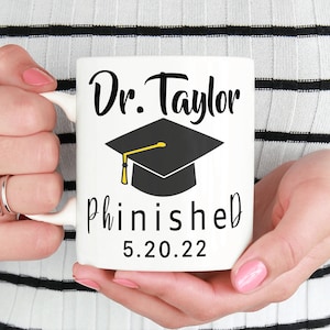 May include: White ceramic mug with black text that reads "Dr. Taylor Phinished 5.20.22". A black graduation cap with a gold tassel is above the text.